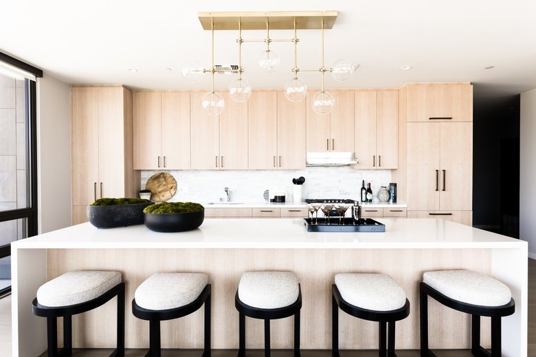 Bar stools at a light wood kitchen island with black bowls of moss and a bra tray. Globe pendant lights above and light wood cabinets.