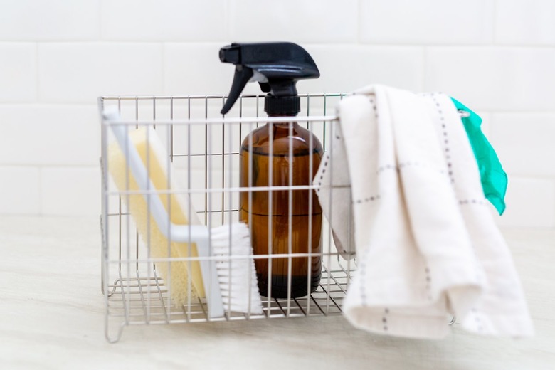 A metal basket with washcloth, gloves, spray bottle, sponge, and brush