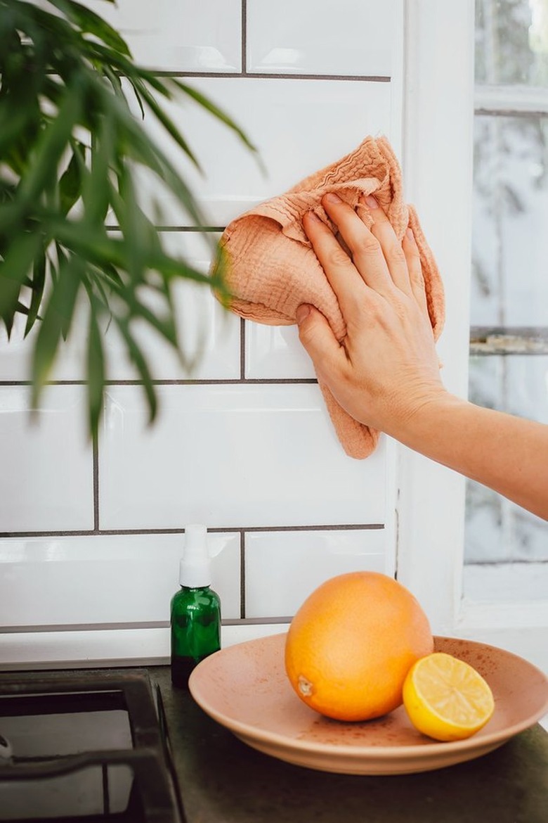 Hand wiping subway backsplash, oranges in wood bowl, plant.