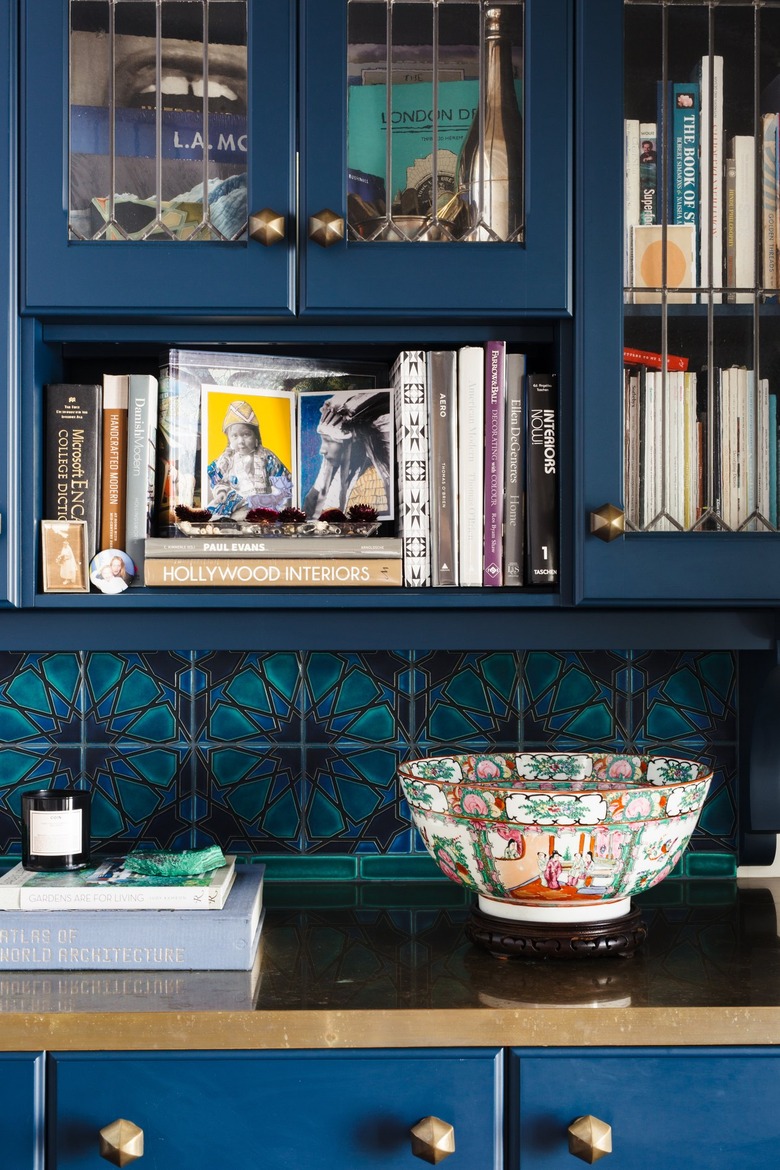 Dark blue kitchen cabinet with gold counter and knobs. Turquoise geometric floral backsplash, vintage bowl and books.