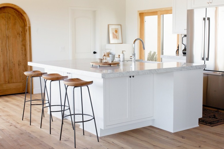 Three bar stools with wooden seats and black metal legs next to a white kitchen island with a marble countertop and chrome faucet. Behind the island is a chrome fridge.