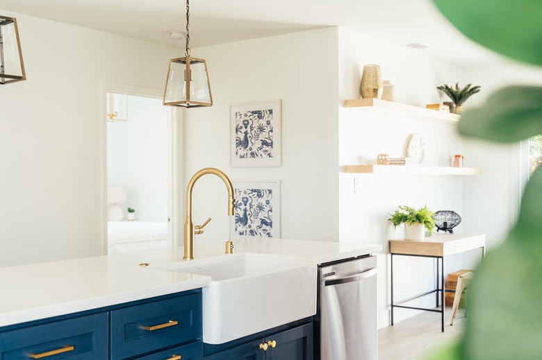 Dark blue kitchen island cabinets with gold hardware, white countertop, and gold sink faucet. Gold lantern pendant lights and wood shelves.