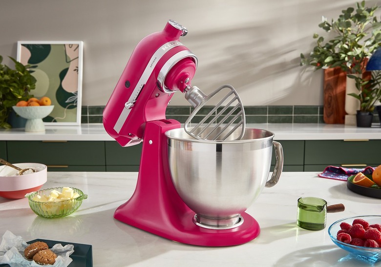 A Hibiscus KitchedAid Mixer rests on a kitchen counter, that also has food and kitchen tools on it, including a bowl of raspberries, a dish of butter, baked goods, an espresso shot filled with water and a plate of oranges. There is another kitchen countertop behind it, with green tiles and a white bowl of oranges, plants, cutting boards and an abstract piece of preen and pink art.