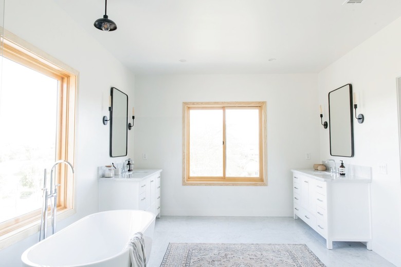 A bathroom with a light wood frame window. Double white vanities, black incandescent light sconces, black frame mirrors are on either side. There is a freestanding tub and a neutral patterned rug.