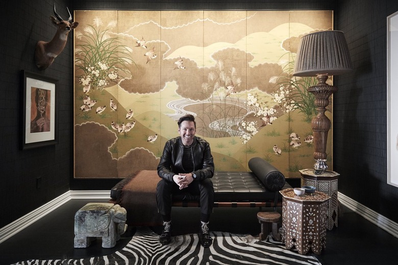 Man with short brown hair is smiling, wearing all black, sitting on lounge chaise with hands folded with large earthy-toned scene panels behind him. Black walls on both sides with other decor in the room, including side tables and zebra throw rug