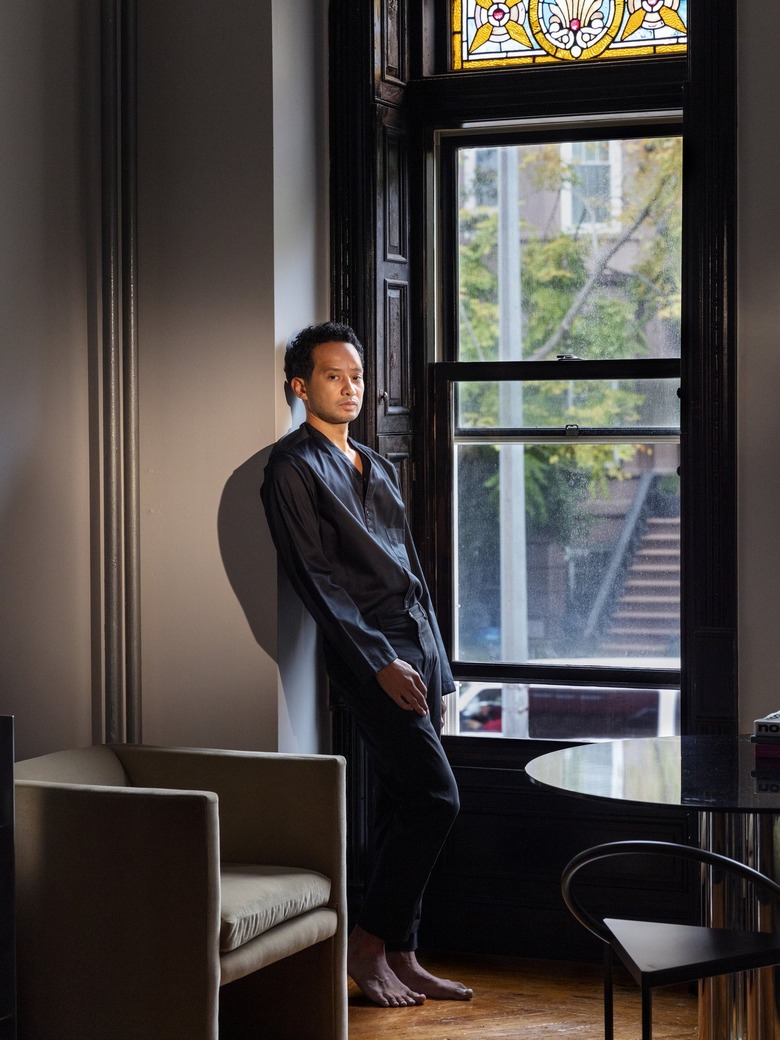 Man with short dark brown hair wearing all black leans against a corner by a window in a beige room with chairs and a table. Outside the window is a brownstone building and staircase.