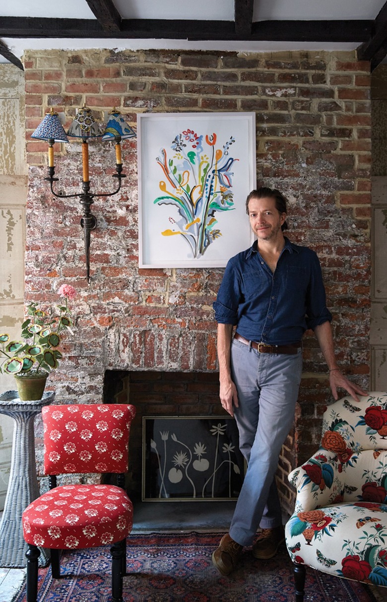 Man with short brown hair grinning and standing up against brick fireplace with legs crossed. He is wearing a blue shirt with rolled-up sleeves and gray pants. There's floral print above the fireplace and an upholstered chair on each side of him.