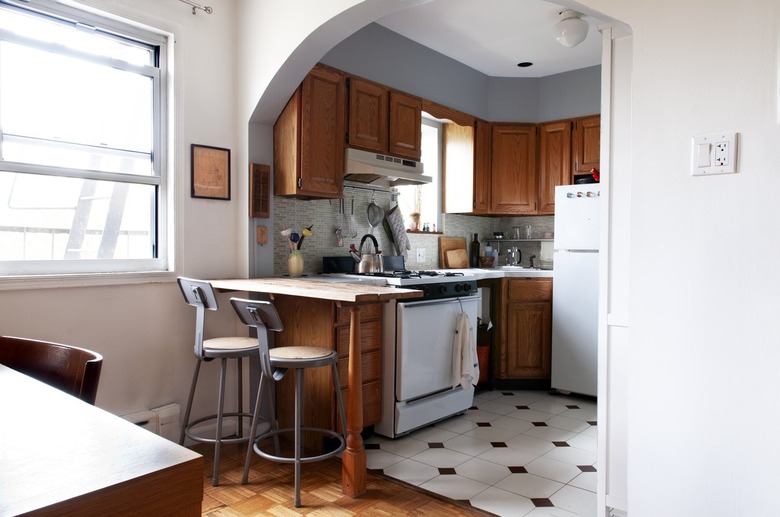 Wood cabinet kitchen with white counters, gray tile backsplash, and metal chair stools, brown-white tile floor.