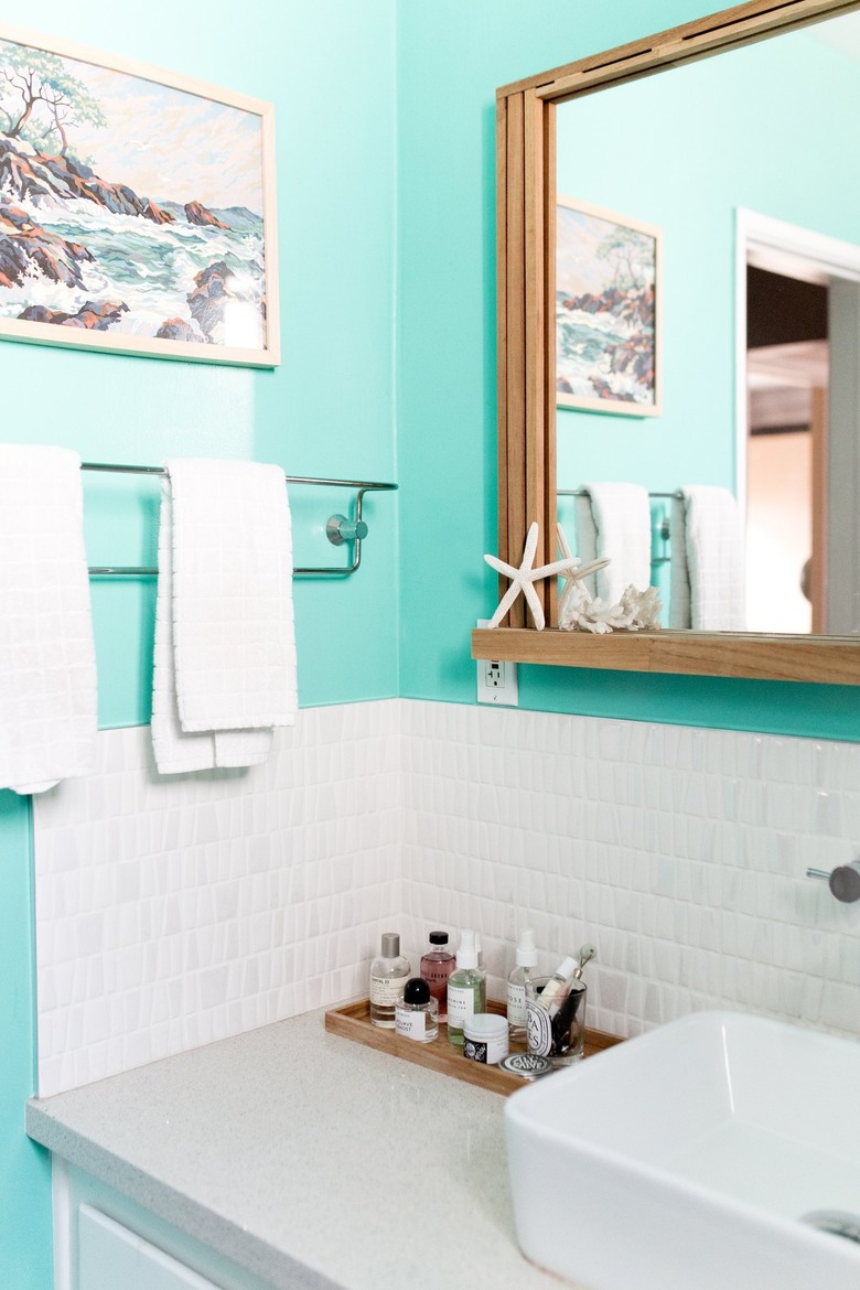 Makeup Organizer Ideas in Bathroom with blue walls, white tile, mirror, towel rack, tray with lotions, make up, painting, white sink.