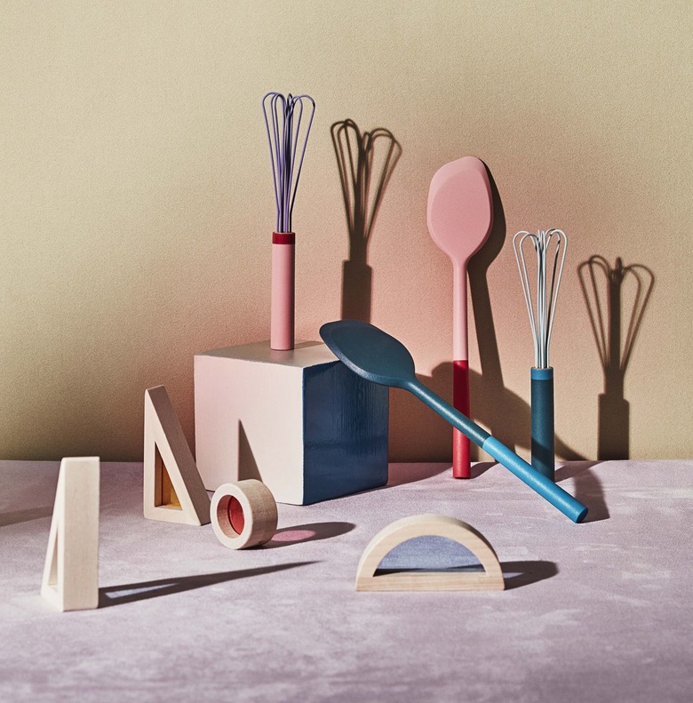 Material's Kids Collection for the kitchen, featuring a mini pink spatula and whisk, along with a mini blue spatula and whisk. These items are surrounded by blocks in fun shapes on a purple marble counter. The background shows orange in the center as it fades to beige around the edges.