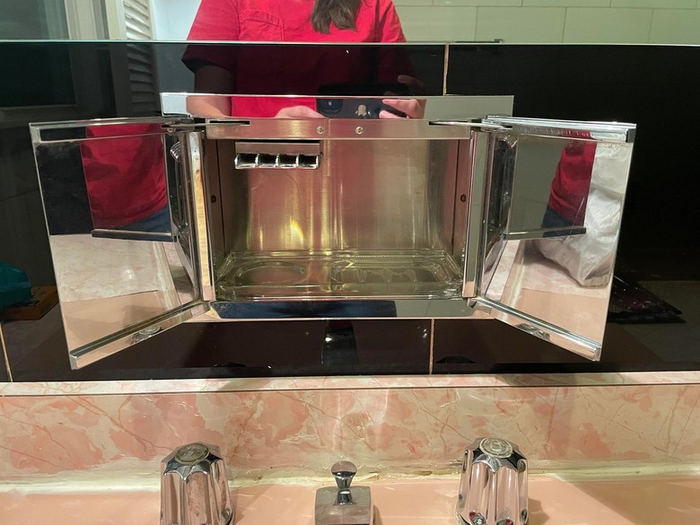 A tiny mirrored storage cupboard beneath a bathroom mirror and above a pink piled sink.