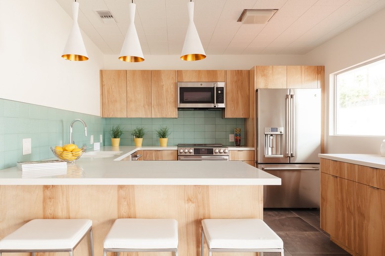 Light wood kitchen cabinets and green backsplash. Yellow planters decorate the counters. Fluted pendant lights and white square stools.