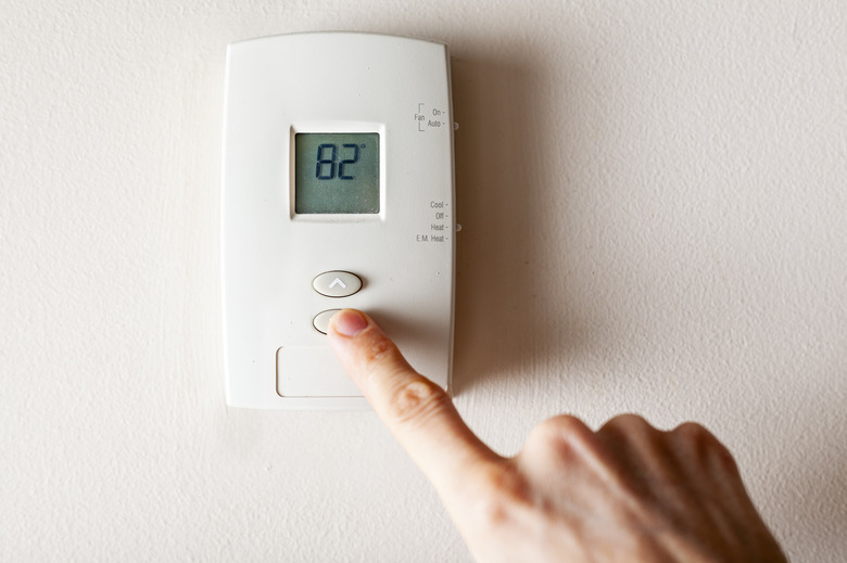 A woman is pressing the down button of a wall attached house thermostat with digital display showing the temperature