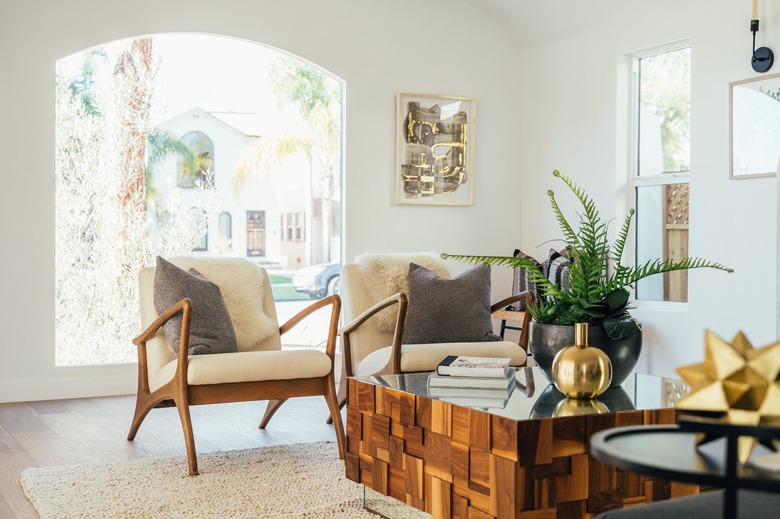 Modernist armchairs with gray pillows, sheepskin. Mirrored tabletop coffee table with wood base, fern plant and gold vase