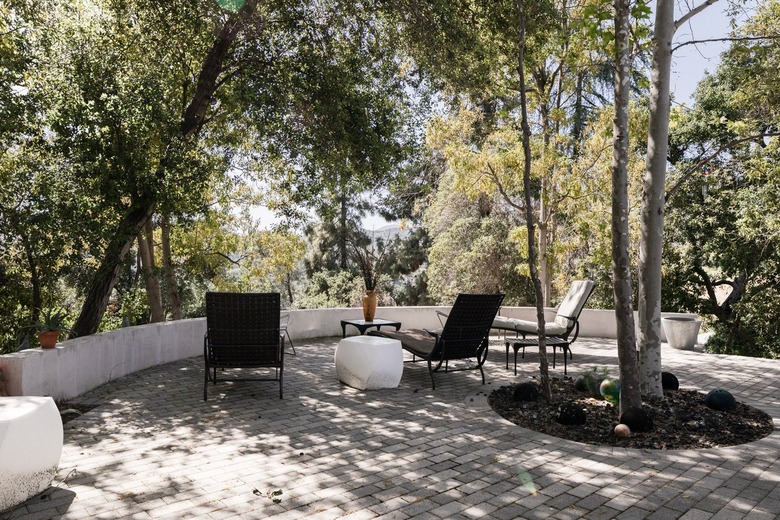 Gray brick patio with chaise lounge chairs, and a white geometric side table. Trees and plants surround.