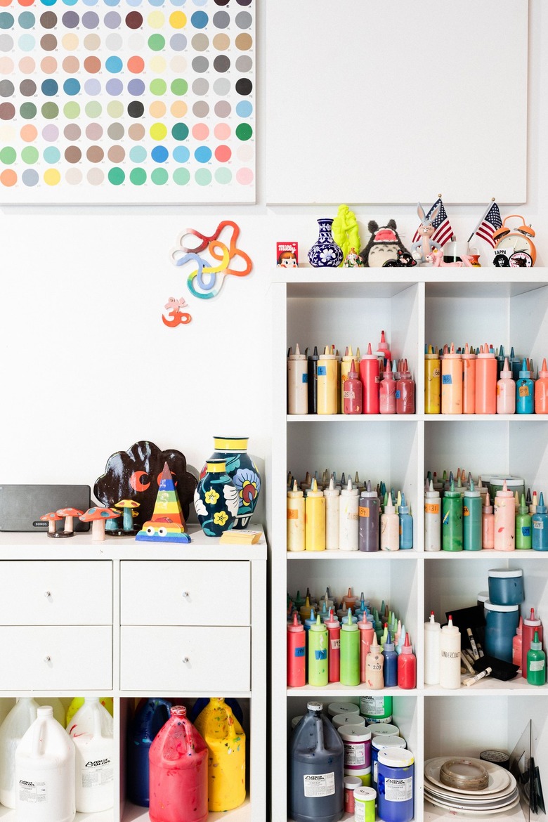 White shelving unit next to small cabinet unit, filled with colored paint bottles, knick-knacks, sculptures, and art supplies against white wall with palette from color wheel