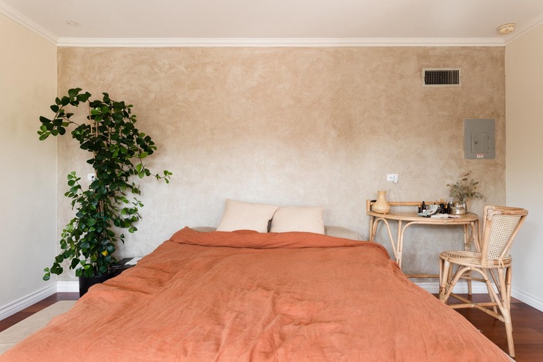 Bedroom with a texture beige accent wall, orange bedding, Boho rattan desk and chair set, and a plant on a trellis.