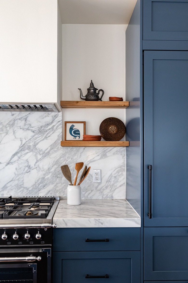 Dark blue kitchen cabinets, marble counter and backsplash, gas range, open shelves.