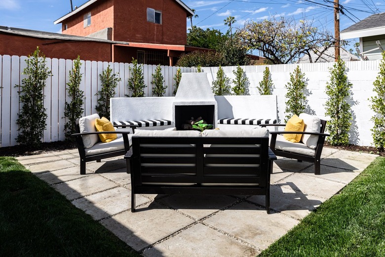 a paver patio with black and white outdoor patio chairs, a white privacy fence with evenly spaced small green trees