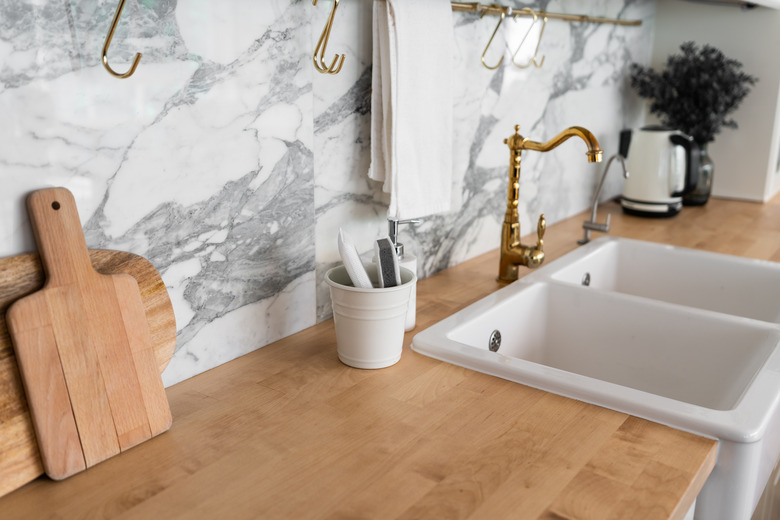 Modern classic kitchen interior with kitchen appliances and white ceramic sink with gold mirror faucet on wood top with marble wall in the background / kitchen interior