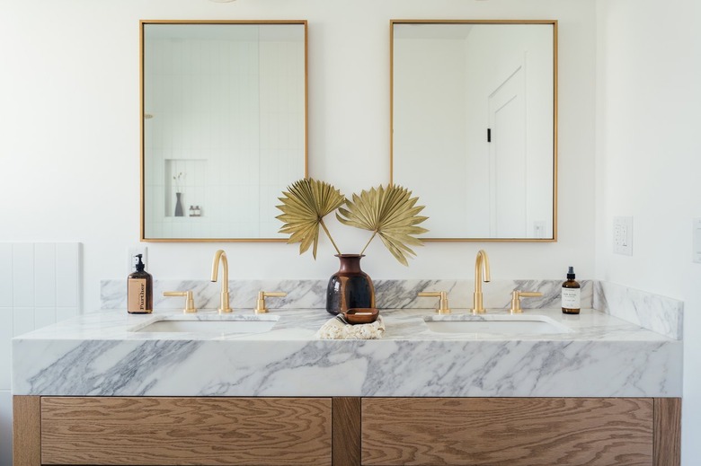 Bathroom sink with white and gray countertop, brass or gold faucet, and mirror. Vase with dried plant.