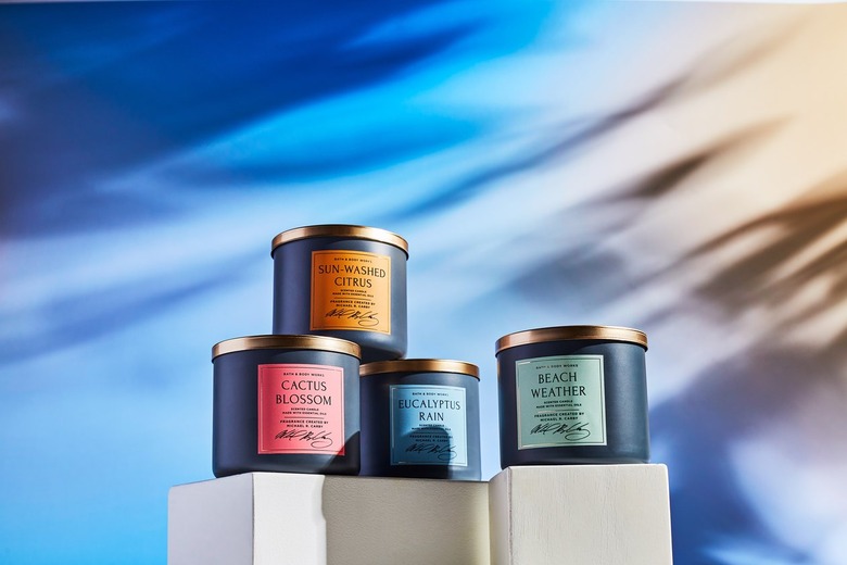 Four Bath and Body Works candles rest on a white cube, against a dark blue and yellow sunlit background. The candles are black with different colored labels and gold tops. From left to right, the candles are: Cactus Blossom, Sun-washed Citrus, Eucalyptus Rain and Beach Weather.