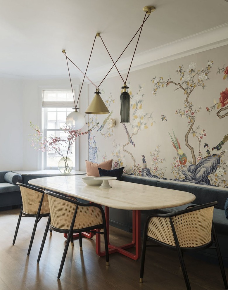 Dining room with chandelier of hanging objects above an oval table with three woven chairs and teal banquette against a wall with floral wallpaper.