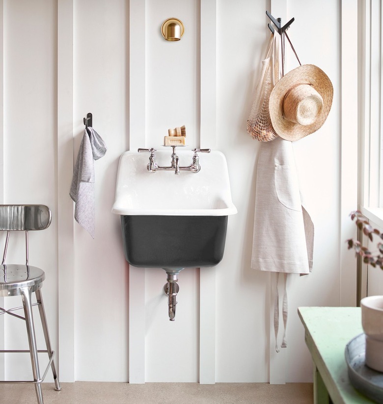 A black cast-iron wall-mounted sink on a white wall with paneling; a hat and an apron are hung near it