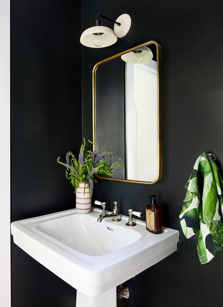 black bathroom with pedestal sink and white sconce above brass mirror