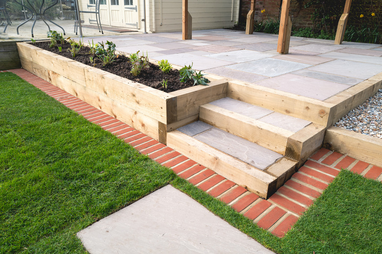 New steps in a garden or back yard leading to a raised patio, alongside a new raised flowerbed made using wooden sleepers. A mowing strip of bricks is in front of newly laid turf.