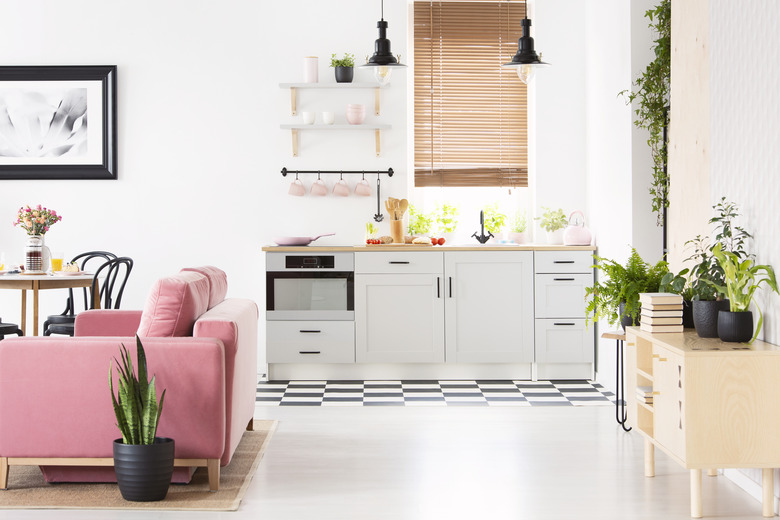 Real photo of open space kitchen interior with checkerboard floor, window with wooden blinds, pink velvet couch and many fresh plants