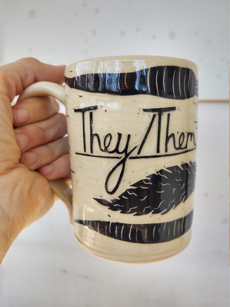 person holding mug with black design that reads "they/them"