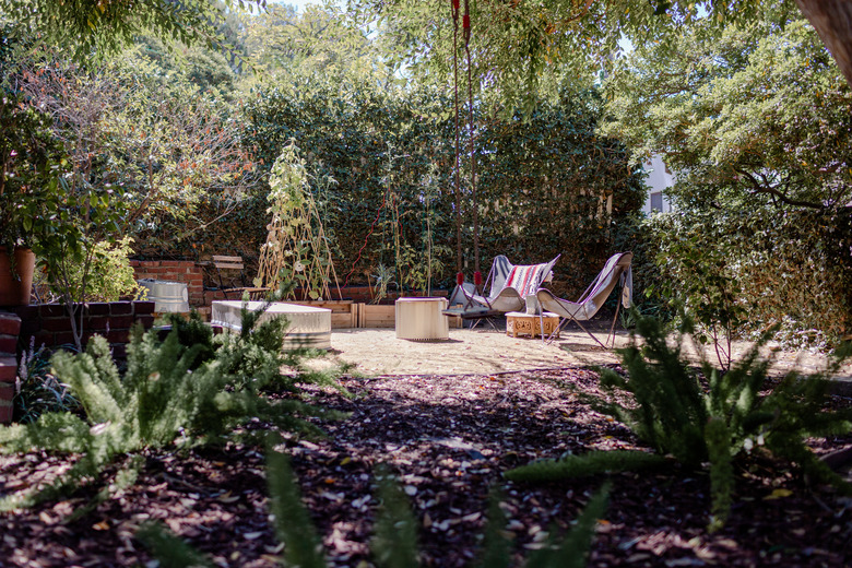 A garden with mulch and a few green plants, a patio with chairs and a table, a fence surrounded by lush trees and shrubs