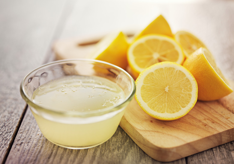 Small glass bowl of lemon juice and several lemon halves arranged on a wooden cutting board