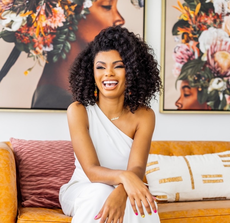 Amber Guyton in a white jumpsuit posing on a burnt orange couch with a rust pillow