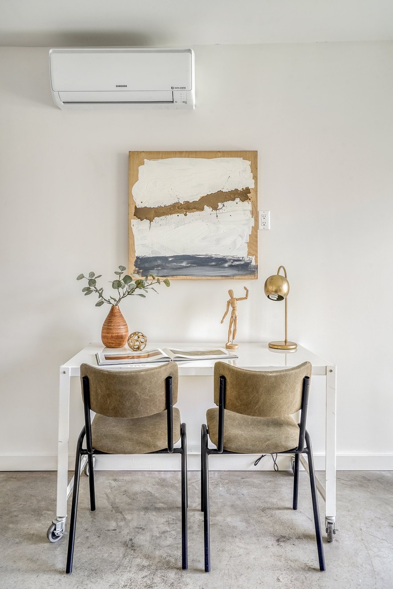 Two neutral-black chairs at a white desk. A vase with plant cuttings, a drawing figurine, a gold globe table lamp, and an abstract painting.