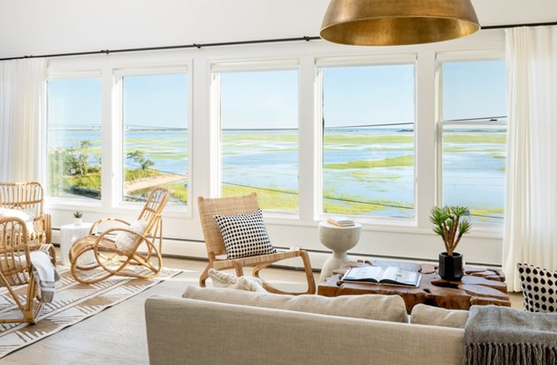 coastal living room with wicker rocking chair, cane lounge chairs, beige couch, brass pendant lamp, large windows with sheer curtains.