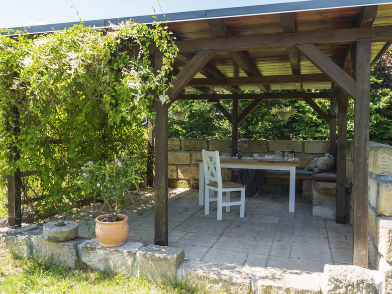 Cozy brown tiber wooden gazebo, pergola with white table, chairs and bench with pillows and blanket surrounded by sandstone wall and climbing plants and blooming pink potted oleander. Sunny summer day.