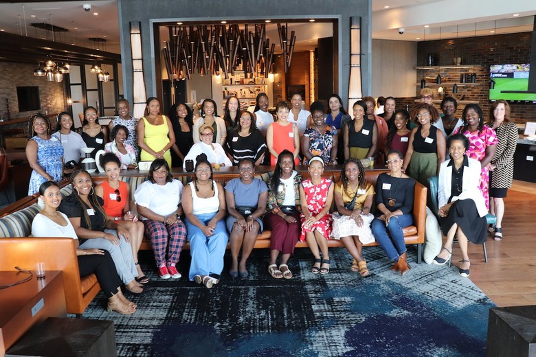 A wide shot of a group of Black women gathered in a hotel lobby. They are sitting on a striped L-shaped couch that