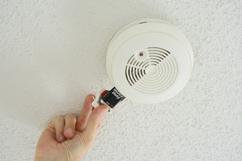 A person replacing the batteries in a white smoke alarm on the ceiling.