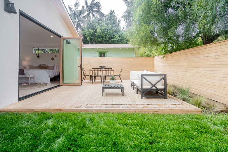 A bedroom with folding glass doors opens up to a backyard with a wood patio, wood horizontal fence, a table with five black chairs and a sofa with a coffee table; green grass and various green plants are planted