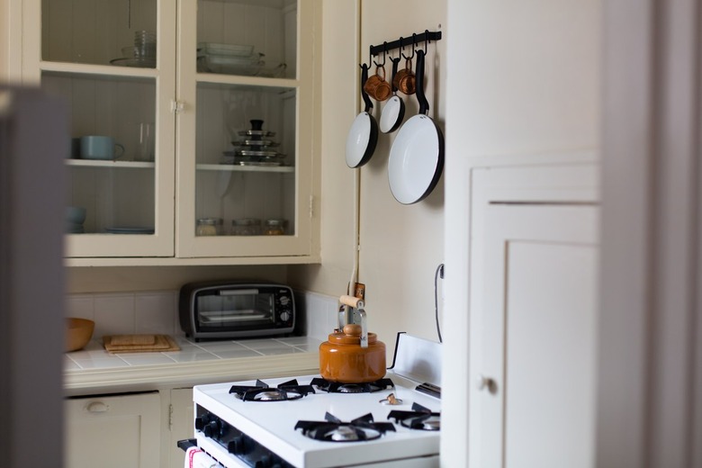 White kitchen with stove and pots and pans hanging on wall with windowed cabinets