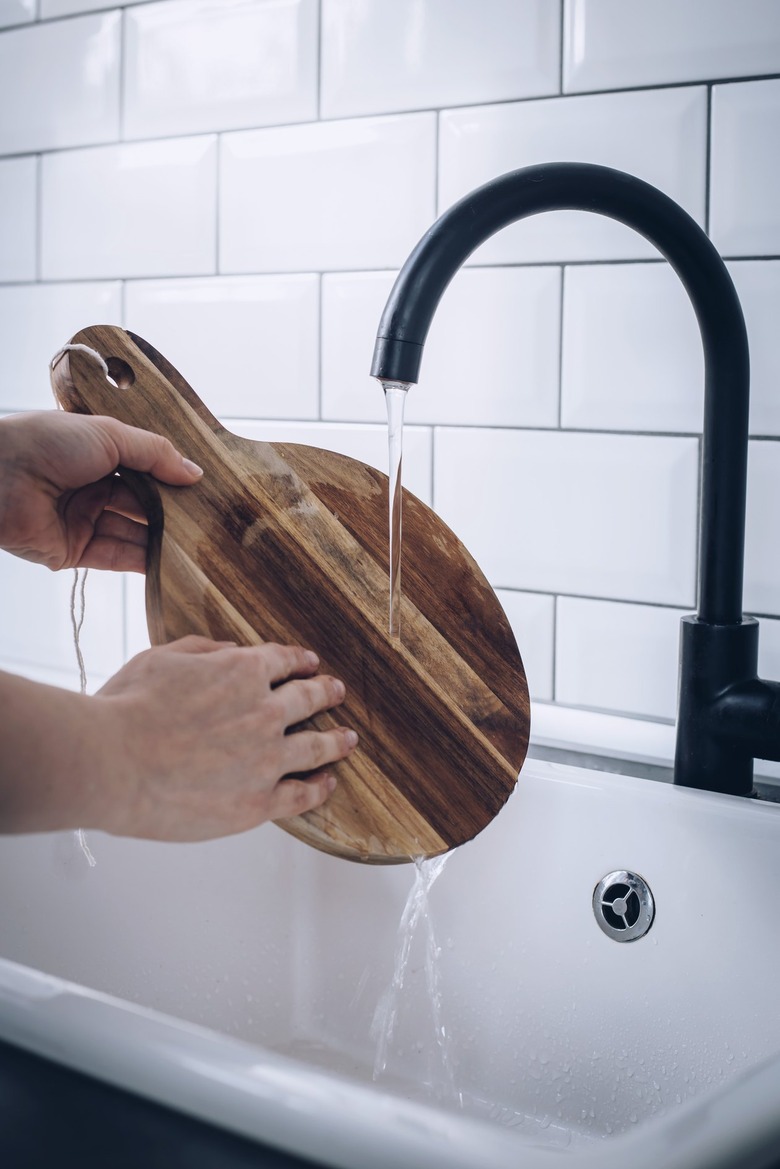 Hands washing round wood cutting board in white sink with black faucet against white tiled backsplash