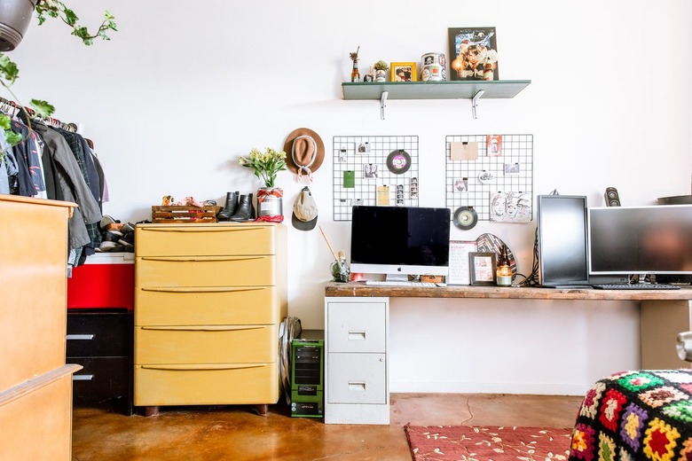 Computers on a desk with a wood tabletop and white filing cabinets. Yellow cabinet, beside. Wire picture boards, hats, and plants as decor.