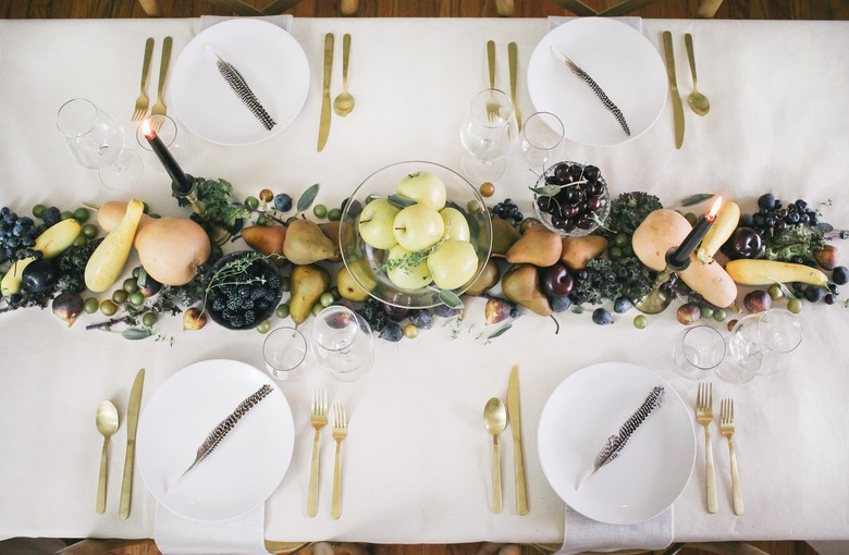 a line of produce, including grapes, pears, cherries, and squash runs down the length of a table with a white cloth and gold cutlery