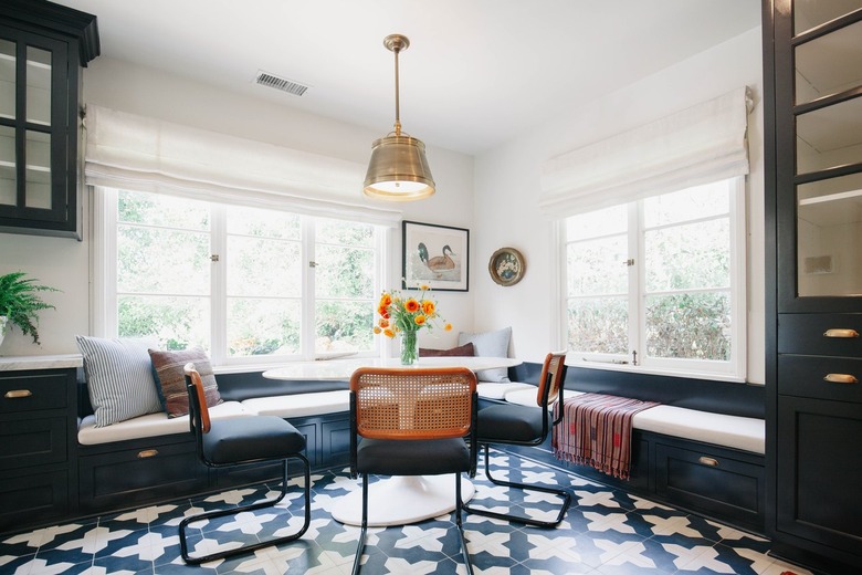 Dining room with white cushioned bench, white flute table with orange tulips, cane chairs, ornate tile floor, and pendant light