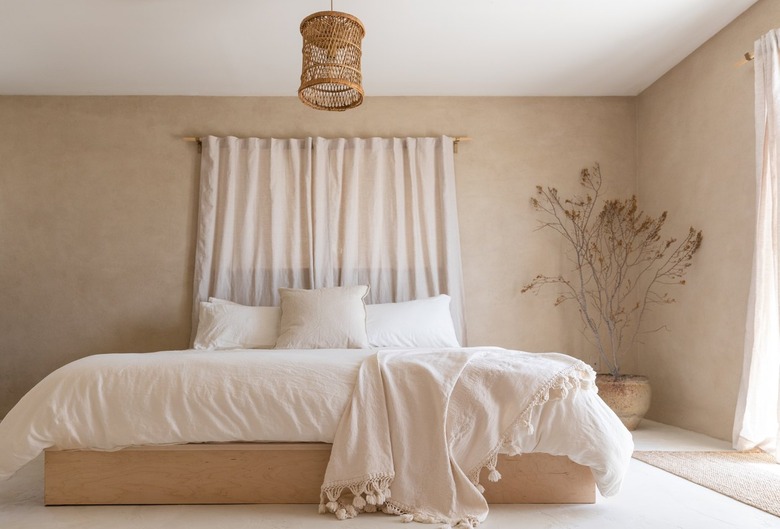 Minimalist bedroom with beige walls, white bedding, tassel blanket, beige curtains, boho pendant light, and dried florals.