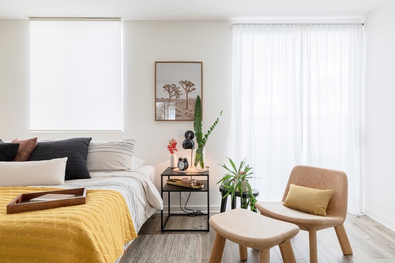 Bedroom with nightstand, beige chair, yellow-white bedding and plants