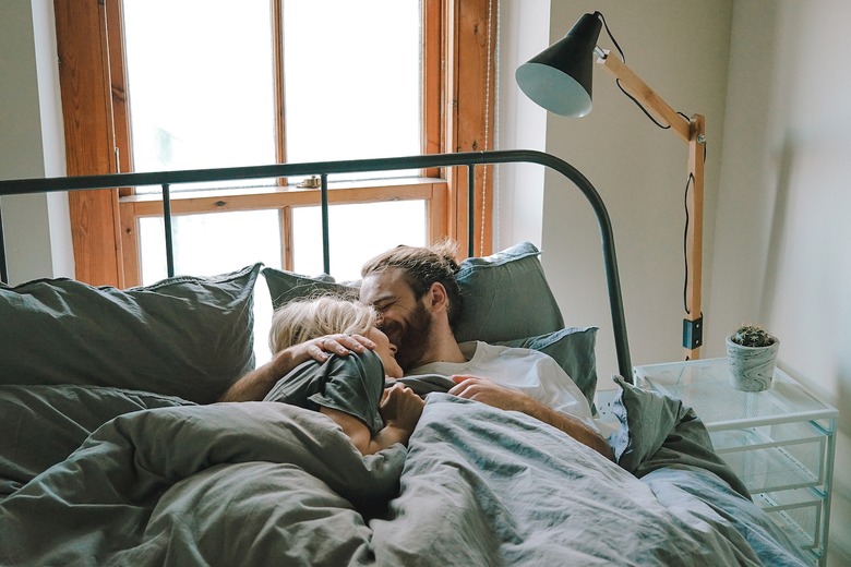 A couple lying in a bed with slate blue sheets next to a clear nightstand and a large floor lamp.