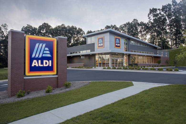 An Aldi sign, surrounded by brick, stands in front of an Aldi store, with trees in the background.